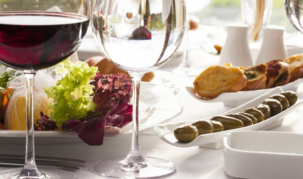 Mesa de reveillon posta, toalha de mesa branca, com petiscos como azeitonas pães e vegetais, com taças vazias e uma taça com vinho.