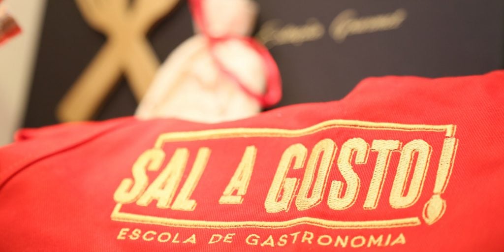 Tecido vermelho com o logotipo da escola de gastronomia Sal a Gosto, fundo descofado com talheres de madeira e totem de chef