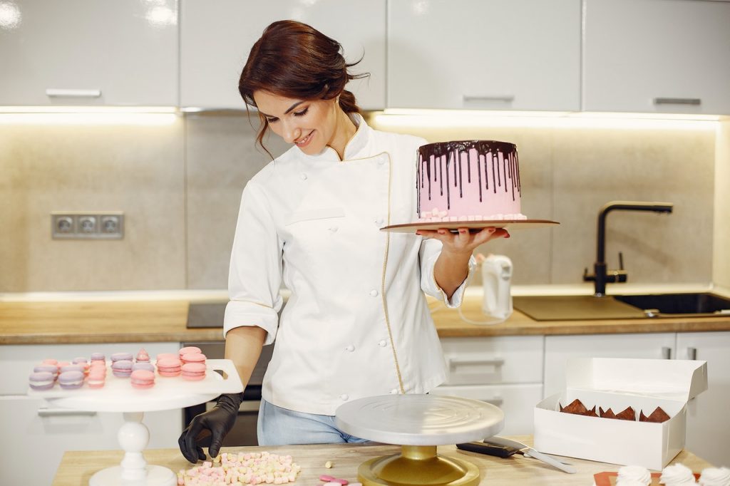 Mulher segurando bolo profissional, com cobertura. Confeitos como macaroons sobre a mesa.
