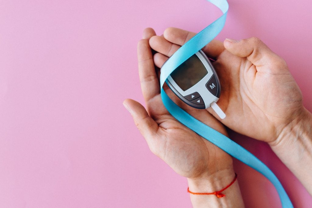 Mãos segurando um medidor de glicose. Com fita azul clara, representando o símbolo da Diabetes, e fundo rosa. É possível registrar os dados obtidos pelo medidor no Aplicativo Diabetes Connect