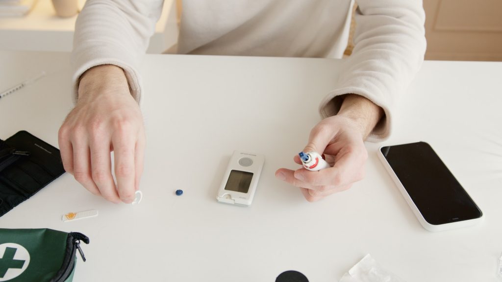 Pessoa prestes a medir a glicose, com todos os aparatos do medidor de glicose sobre a mesa, e um smartphone ao lado, representando como usar o Aplicativo Diabetes Pal