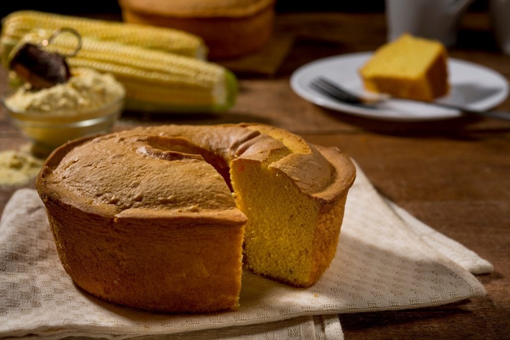 bolo de milho cortado e faltando uma fatia, que está ao fundo da imagem em um prato branco com um garfo do lado; ao fundo também aparecem milhos