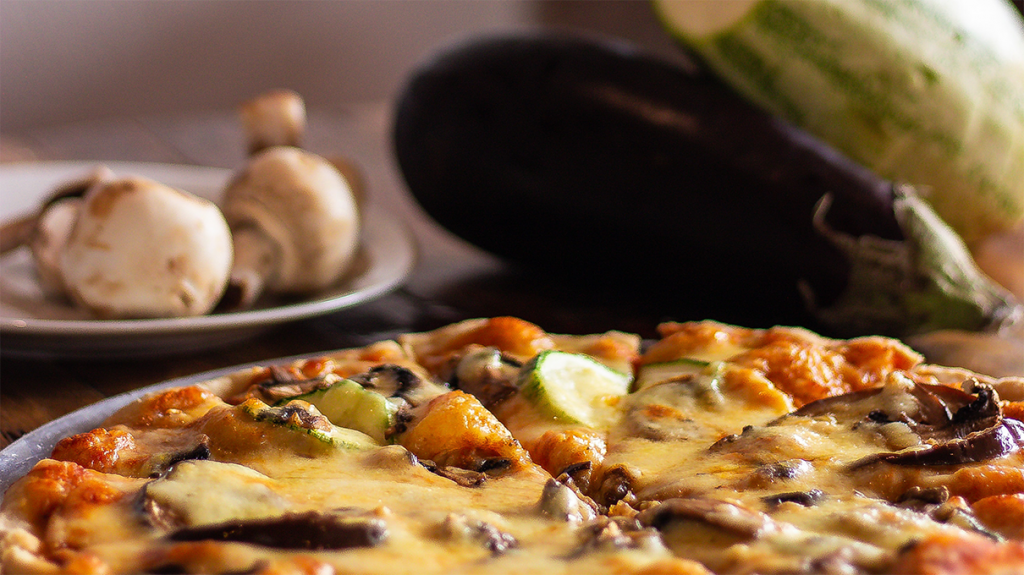 Pizza de beringela com queijo, com cogumelos, beringela e abobrinha ao fundo. Simbolizando Berinjela e receitas