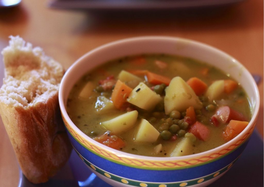 Imagem com uma tigela de sopa de legumes ao lado de um pedaço de pão