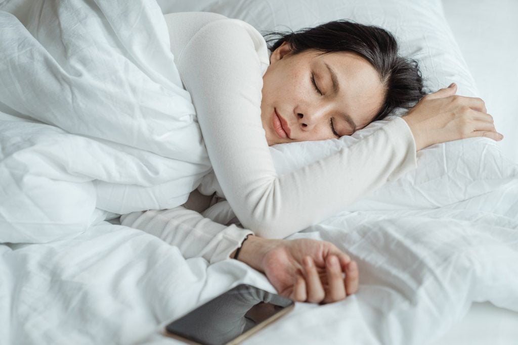 Mulher dormindo, celular com tela apagada próximo à mão dela sobre a cama. Simbolizando a dúvida se é possível emagrecer dormindo
