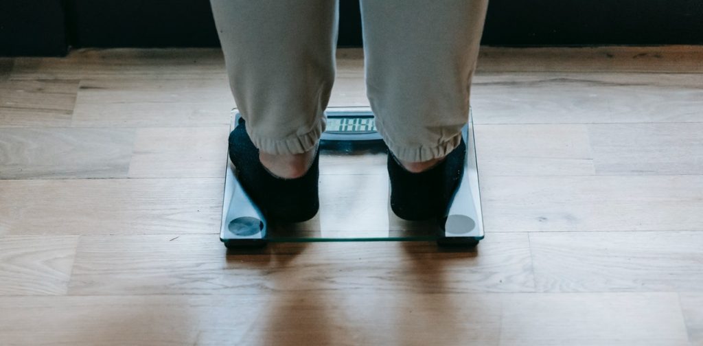 Pessoa de meias pretas e calça moletom sobre balança de medir peso de vidro, simbolizando a dúvida: como saber se meu peso é ideal para minha altura