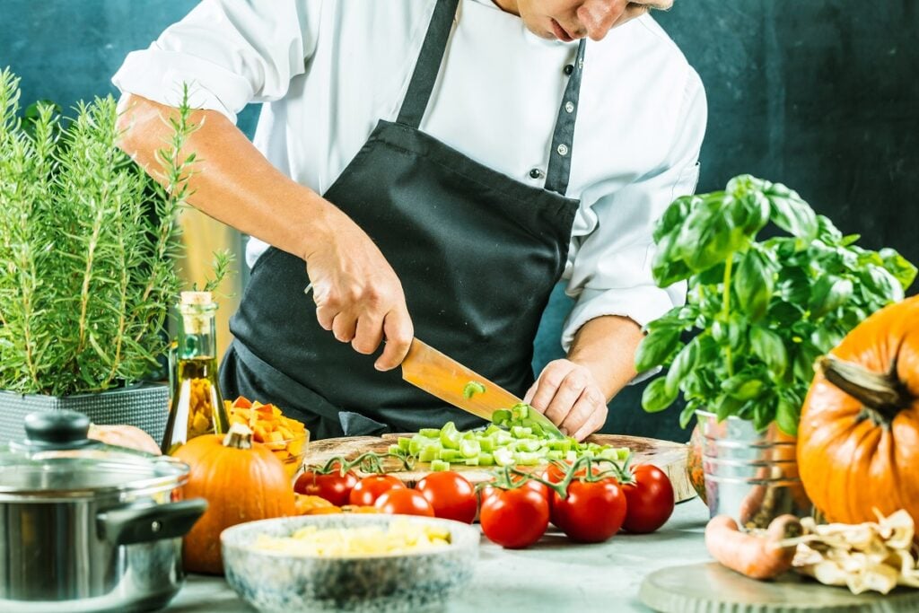 cozinheiro cortando salsao na tábua; na mesa também há tomates e outros vegetais e legumes