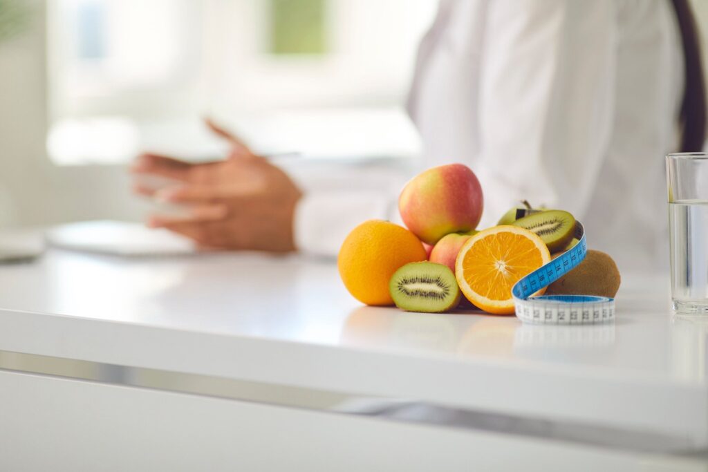 Nutricionista de jaleco branco ao fundo explicando como fazer Dieta para perder até 5kg em 2 semanas. Frutas, fita métrica e copo de água no primeiro plano.