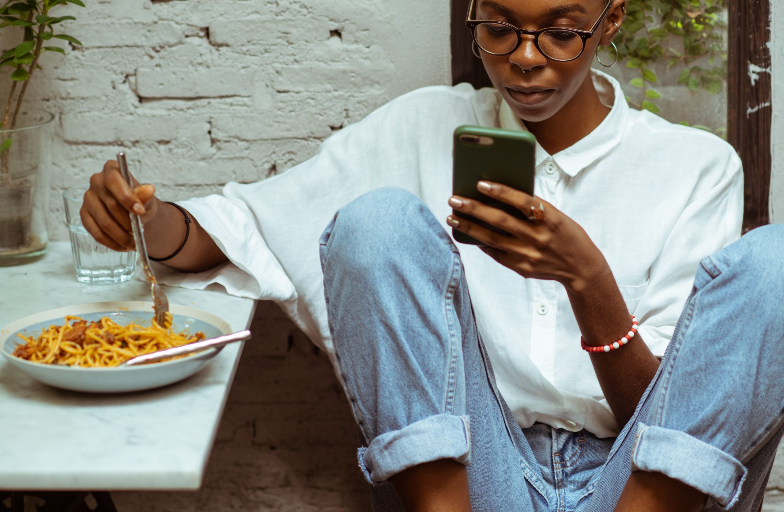 Mulher comendo macarronada enquanto mexe no celular. Distrações como essa devem ser evitadas numa reeducação alimentar