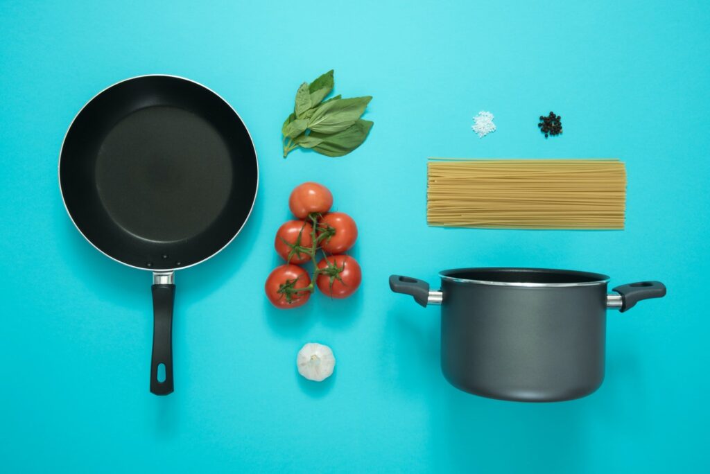 Frigideira, panela funda, massa crua de macarrão, tomates, manjericão, alho, sal e pimenta. Utilizados no Curso Culinária Italiana Completa Escola Sal a gosto. Fundo azul ciano.