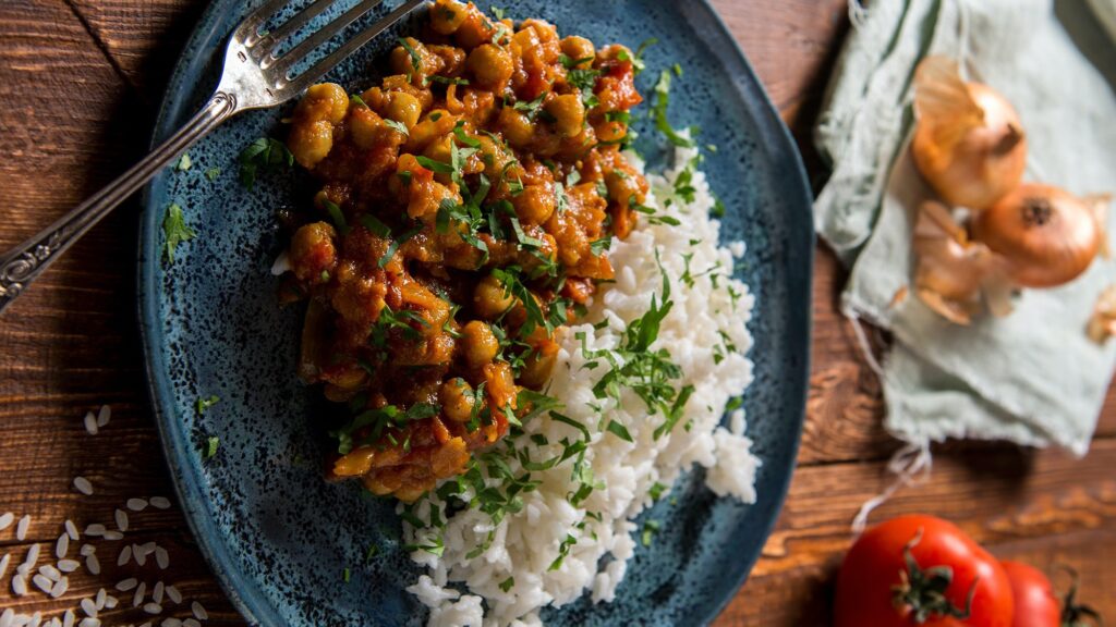 Cebolas e tomate ao fundo. Prato de feijão com arroz soltinho, que são parte do conteúdo do Curso cozinha para iniciantes Eduk