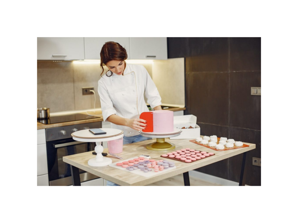 Mulher confeitando bolo profissionalmente, com outros confeitos como macaroons sobre a mesa. Sobre suporte na mesa, um celular (que é um dos dispositivos no qual é possível assistir ao Curso completo Clube da Mah