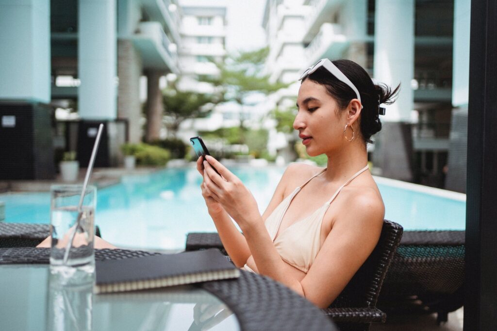 Mulher sentada à mesa usando celular, onde pode ser baixado o Aplicativo Glic. Em ambiente elegante, com piscina. A mulher usa óculos escuros pendurado na cabeça, brinco dourado de argola e biquíni. Livro e copo de água com canudo de metal sobre a mesa.