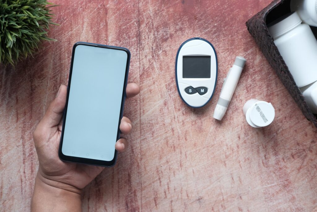 Aparelho para diabetes medidor de glicose, medicação e vaso sobre mesa. Mão segurando celular ao lado, onde pode ser instalado o Aplicativo My Sugr