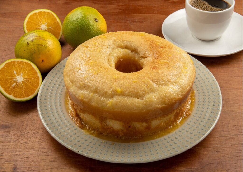 Imagem com um bolo de laranja fofinho ao fundo e algumas laranjas na lateral da foto