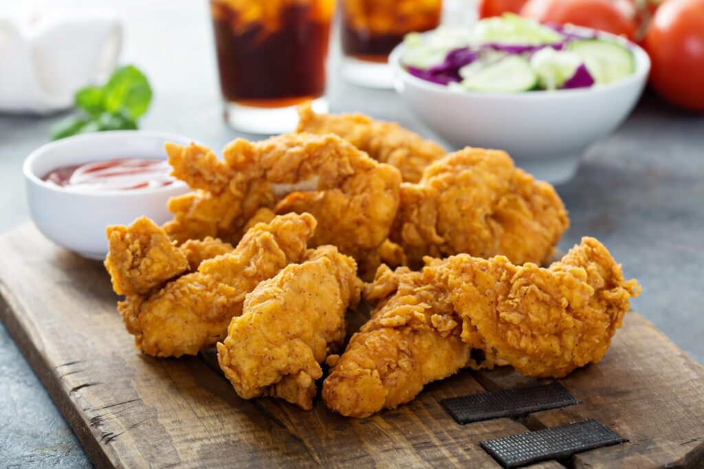 Frango frito em tábua com molho, copo e acompanhamentos ao fundo