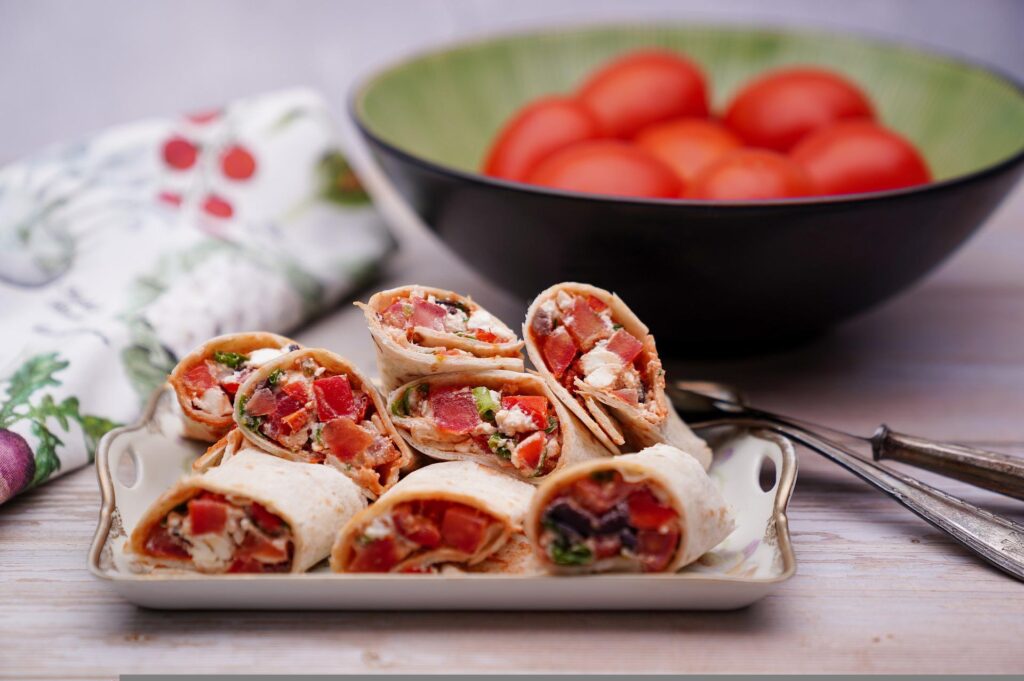 Wrap com recheio de tomate. Talheres, vasilha com tomate ao fundo e pano de prato. Wrap é uma opção de comida leve para almoço