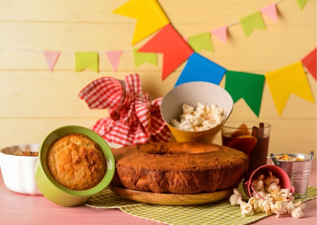 Imagem colorida com bandeiras de são joão ao fundo com diversos pratos típicos de comidas juninas