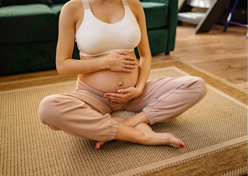 mulher grávida sentada com as pernas cruzadas e mãos na barriga; ela veste um top branco e uma calça creme
