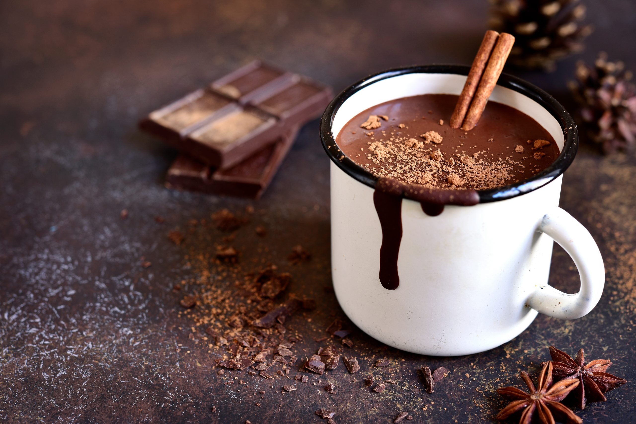Caneca com chocolate quente, nessa receita há canela. Barra de chocolate ao fundo.