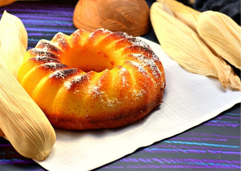 bolo de milho redondo ao lado de algumas espigas em mesa azul