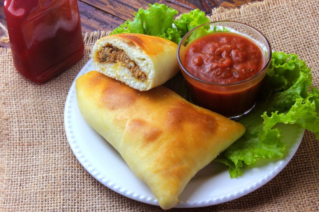 Receita esfiha fechada, um pedaço cortado ao meio mostra recheio de carne. Sob essa esfiha cortada, está uma esfiha fechada no prato. No prato também há alface e um potinho com ketchup. Frasco de ketchup sobre a mesa, ao lado do prato com as duas esfihas.