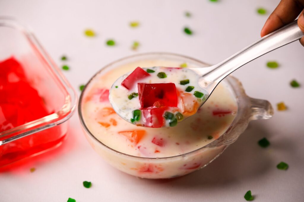 Mão segurando talher, dando uma colheirada em receita de gelatina colorida. Ao lado, uma travessa com gelatina vermelha