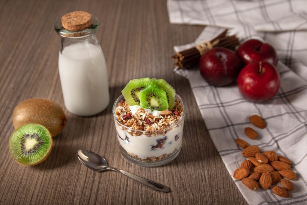 Receita iogurte natural, na garrafa com ele mais líquido e um pote de vidro com a versão cremosa. Acompanha grãos e frutas como kiwi e maça. Toalha de mesa e colher ao redor dos iogurtes sobre a mesa.