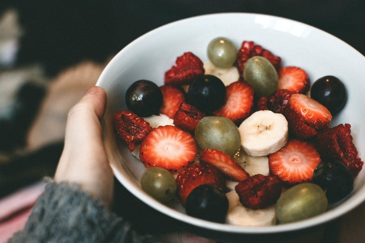 pessoa segurando pote branco de salada de frutas com morango, uvas, banana e mais frutas