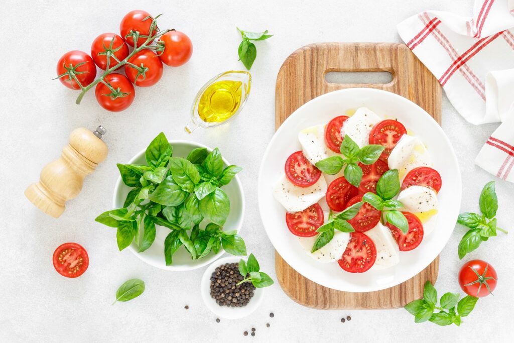 salada caprese servida em prato branco e tábua de madeira; ao lado, os ingredientes inteiros da salada