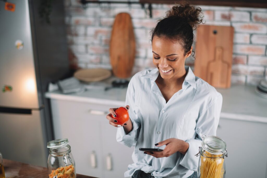 Mulher em uma cozinha segurando um tomate em uma mão e um celular na outra