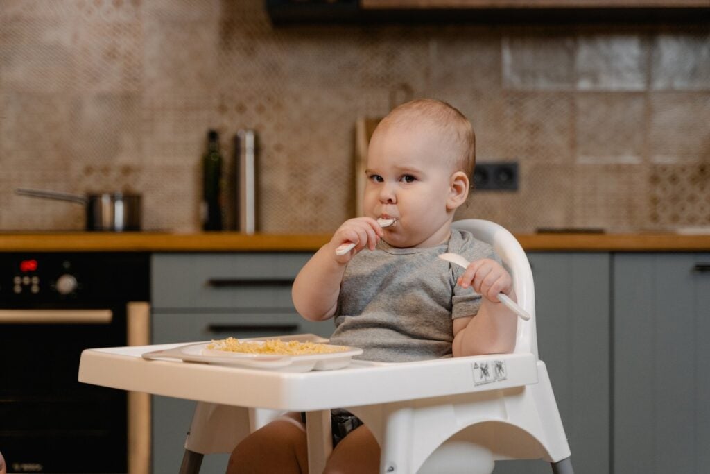 Bebê em cadeirão, em cozinha, comendo um prato com comida. Simbolizando como melhorar a imunidade do bebê com alimentos
