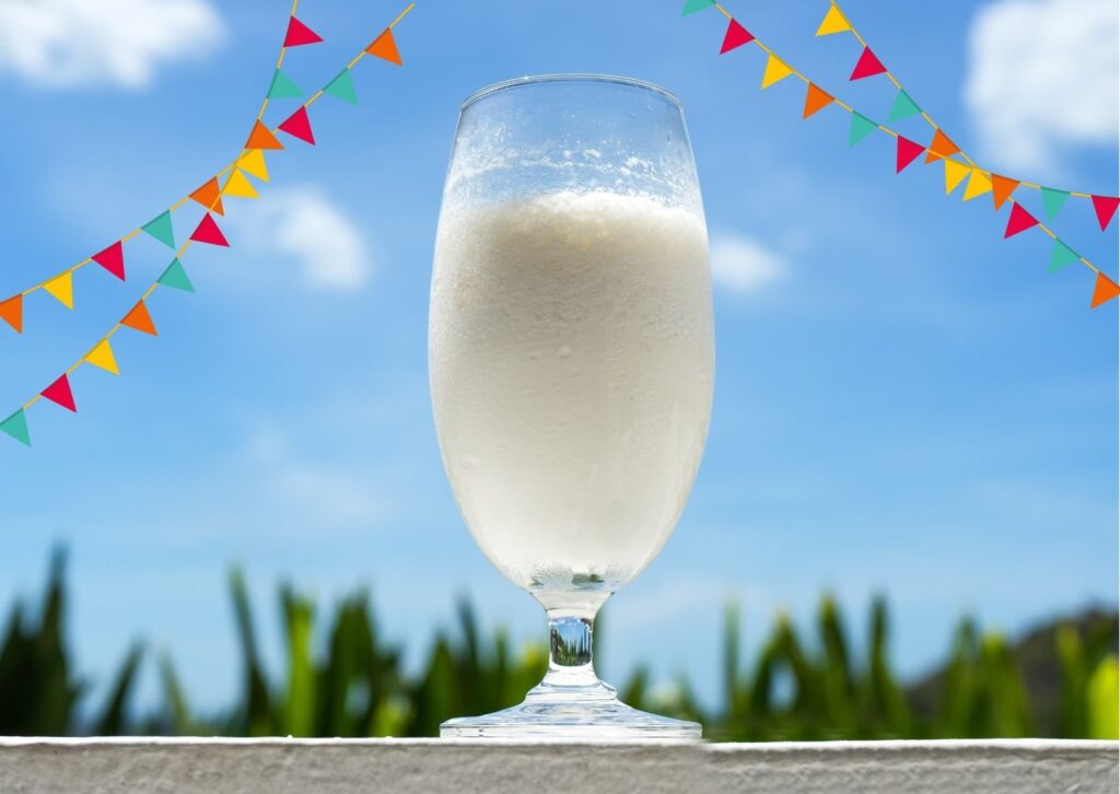preparo da batida de coco em copo transparente; ao fundo, o céu azul com bandeirinhas de festa junina