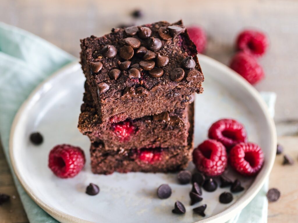 Prato branco ao fundo com três pedaços de brownie e frutas ao lado