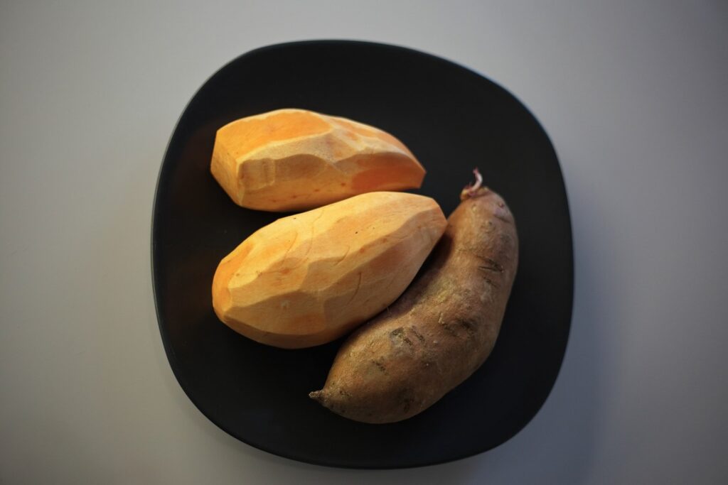 Uma batata doce com casca e duas descascadas dentro de prato. Representando receitas com batata doce