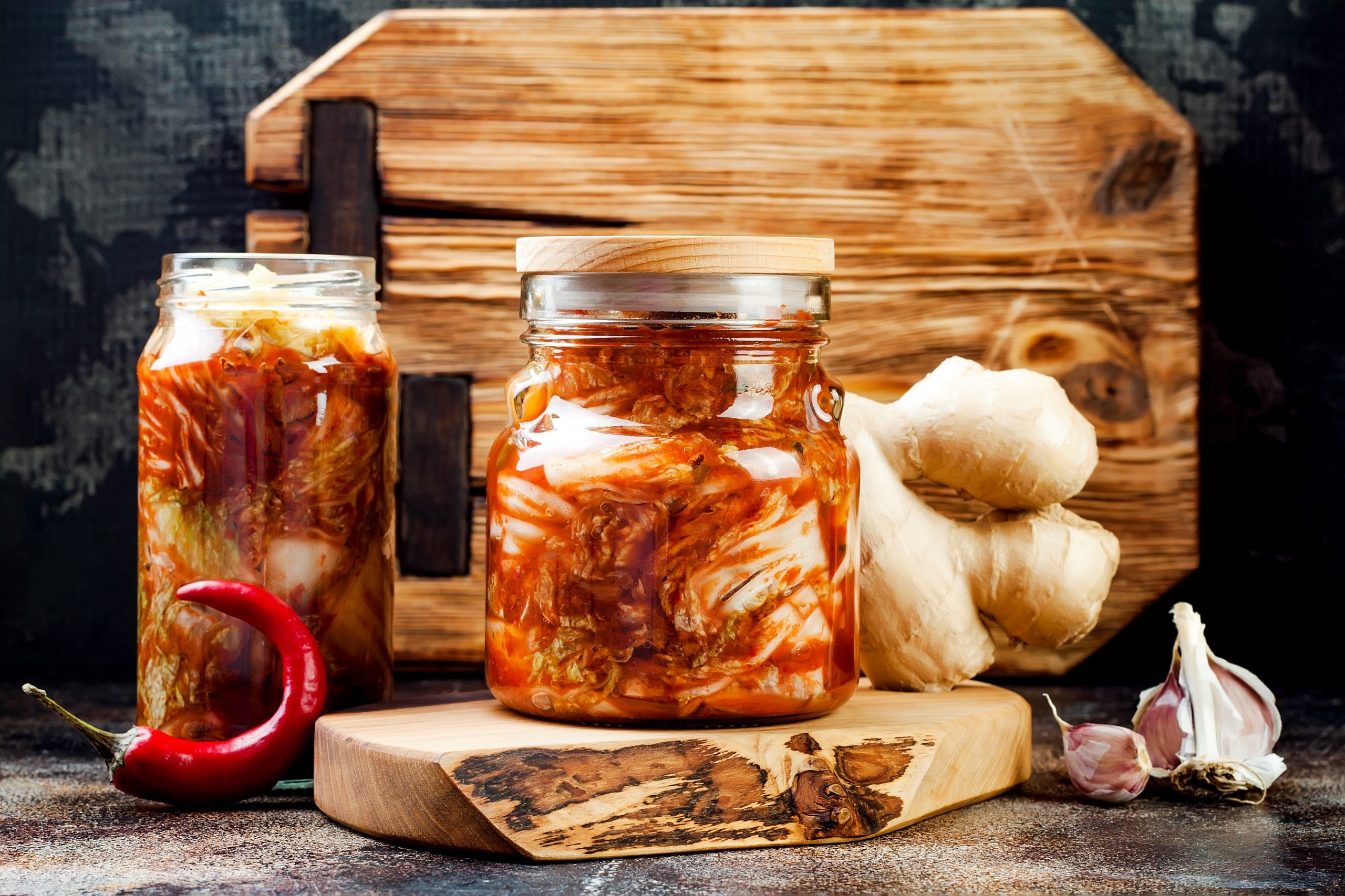 Pote de vidro de kimchi em cima de madeira redonda com pimenta, gengibre e alho ao lado