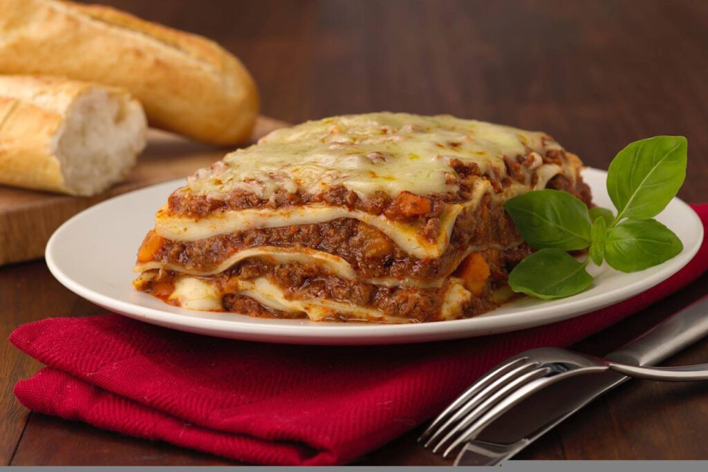 Receita de lasanha no prato com garfo ao lado, pano de prato vermelho sob o prato e baguete de pães ao fundo.