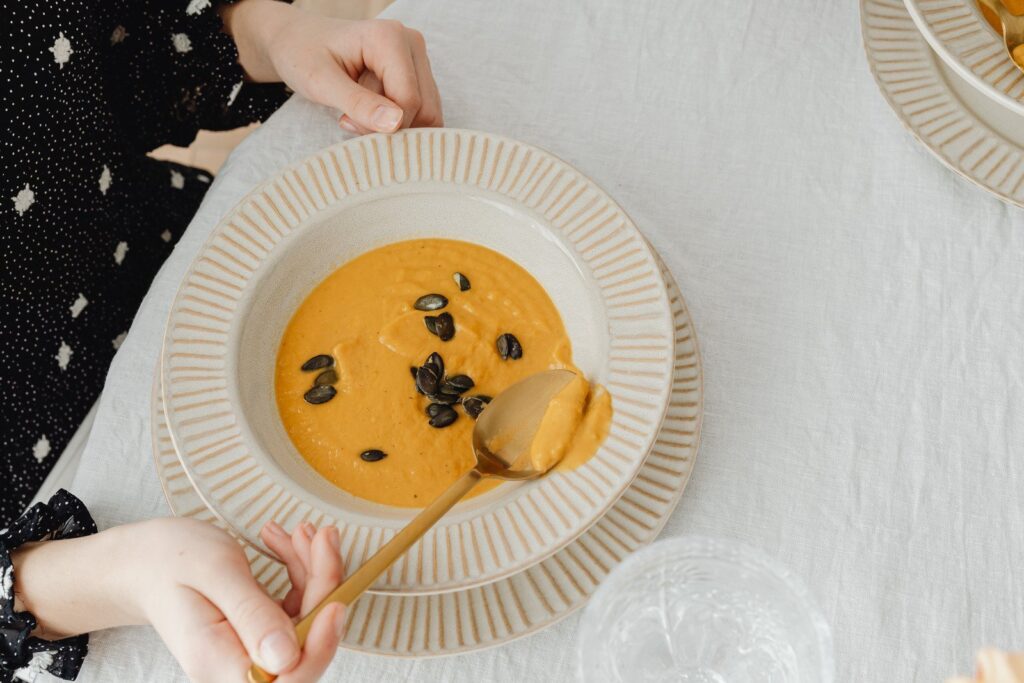 Prato de sopa laranja com semente sobre a mesa. Mulher sentada à mesa pegando uma colher dessa sopa de abóbora, que é uma das opções de sopa seca tudo