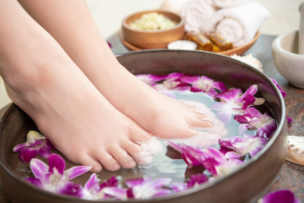 Mulher mergulhando os pés em tina com água e flores no piso de madeira.