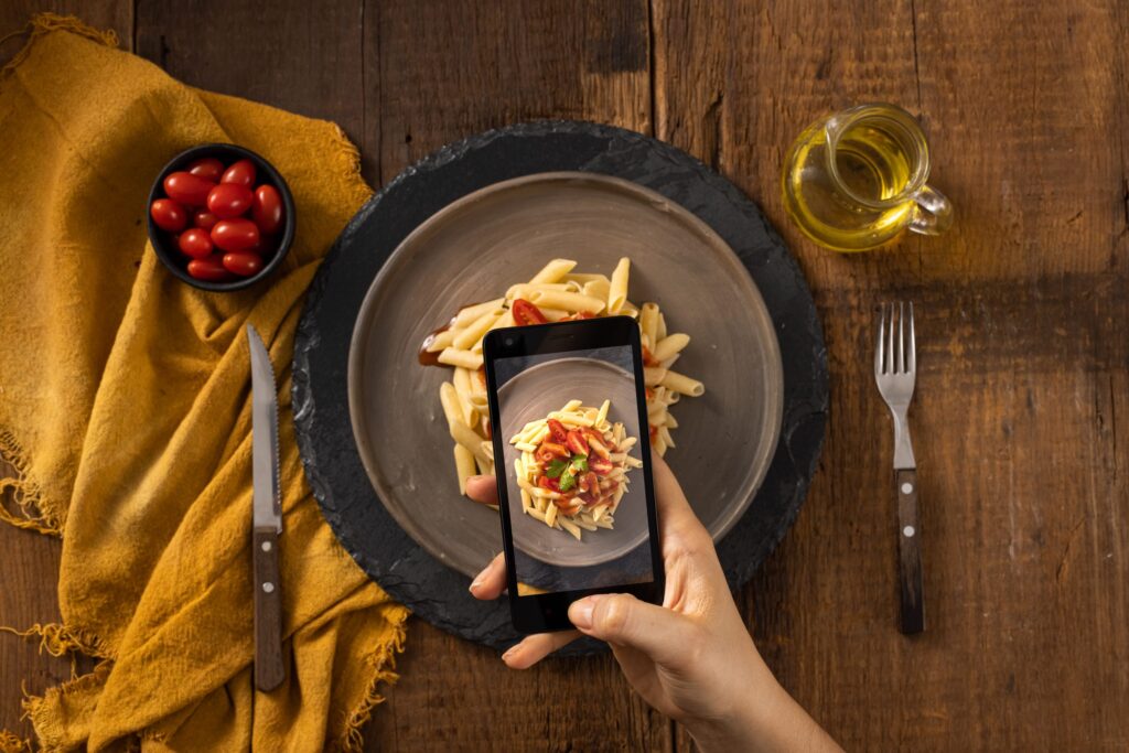 Mão segurando celular em cima de prato de massa com molho de tomates.