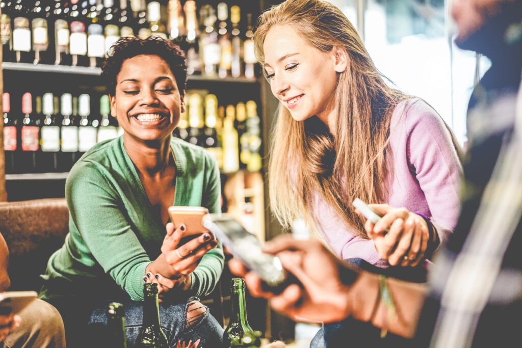Mulheres olhando celular com parede de bebidas ao fundo
