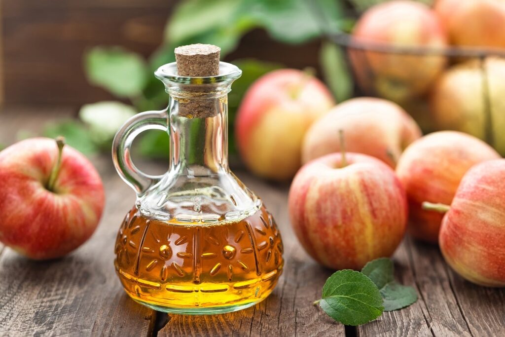 vidro com vinagre de maçã dentro, ao lado de maçãs inteiras em superfície de madeira